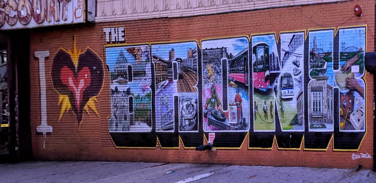 Mural sobre una pared de ladrillo naranja con la palabra BRONX en letras gigantes, cada una rellena con escenas pintadas del barrio —el metro elevado, los edificios, la gente de las esquinas— y precedida de un corazón rojo radiante.