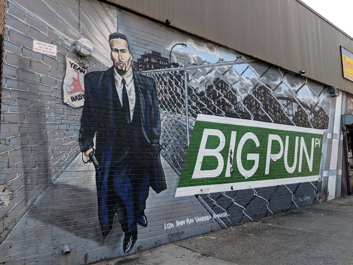 Mural de Big Pun sobre una pared de ladrillo gris: el rapero, de traje oscuro, camina hacia el espectador junto a una placa de calle verde que dice «BIG PUN PL», con una valla metálica y el perfil de la ciudad pintados al fondo.