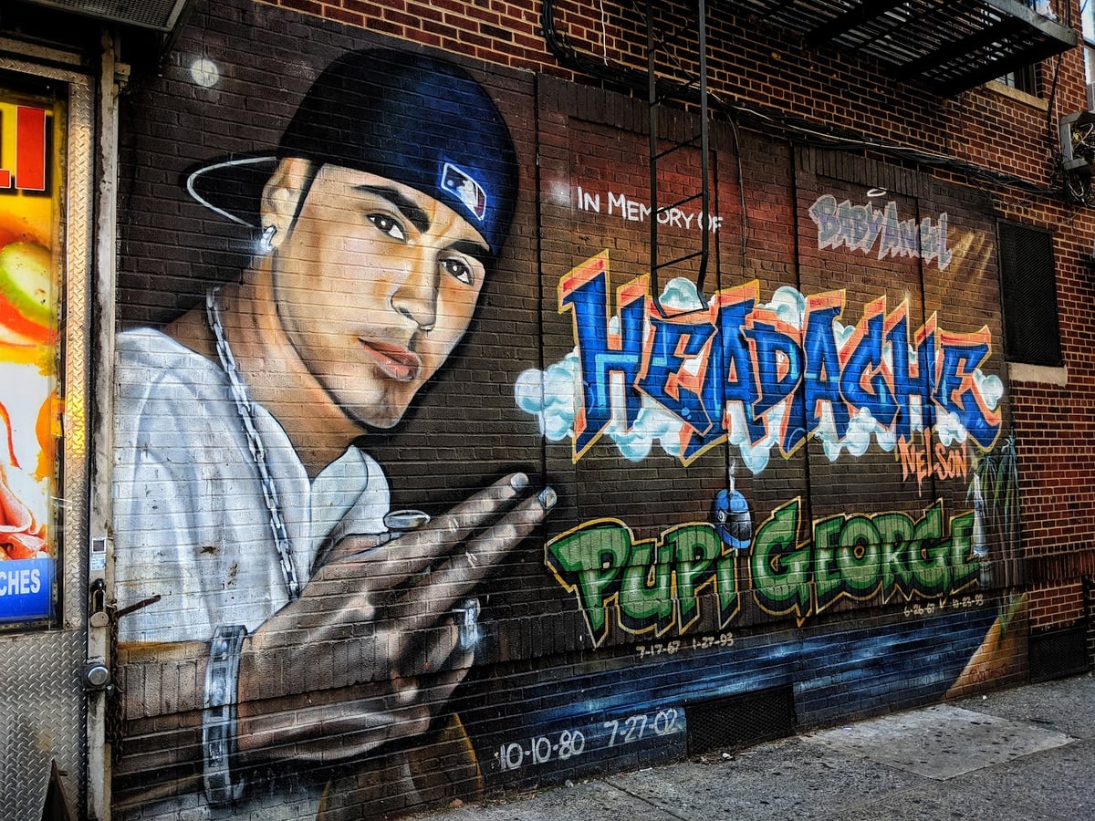 Mural conmemorativo con el retrato de un joven con gorra de béisbol y cadena al cuello, junto a los nombres «Headache» y «Pupi George» en letras de grafiti, la leyenda «In memory of» y las fechas de nacimiento y muerte de ambos.