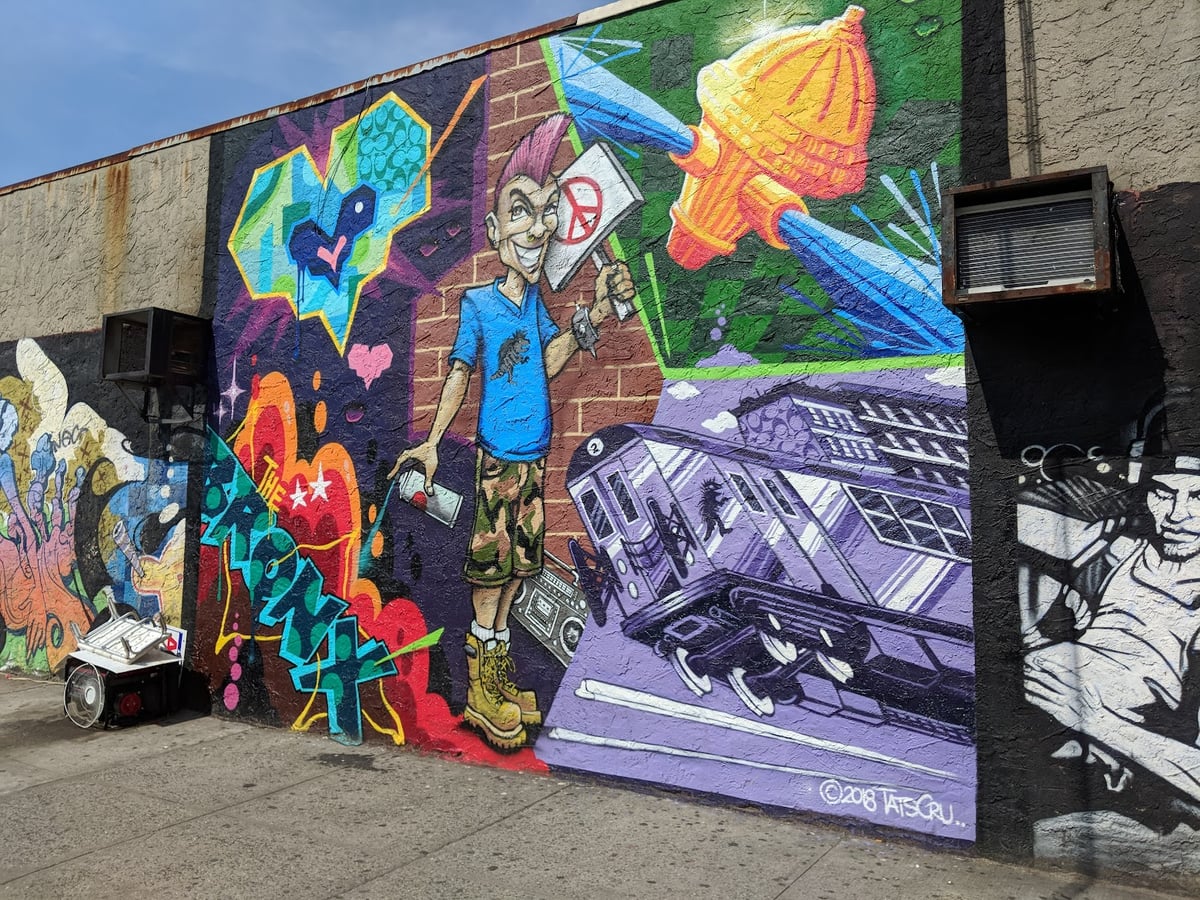 Mural firmado por TATS CRU en 2018, con un chaval de cresta rosa y un bote de spray en la mano, un vagón de metro en tonos morados, un corazón y las letras de grafiti «The Bronx» sobre un fondo de colores intensos.