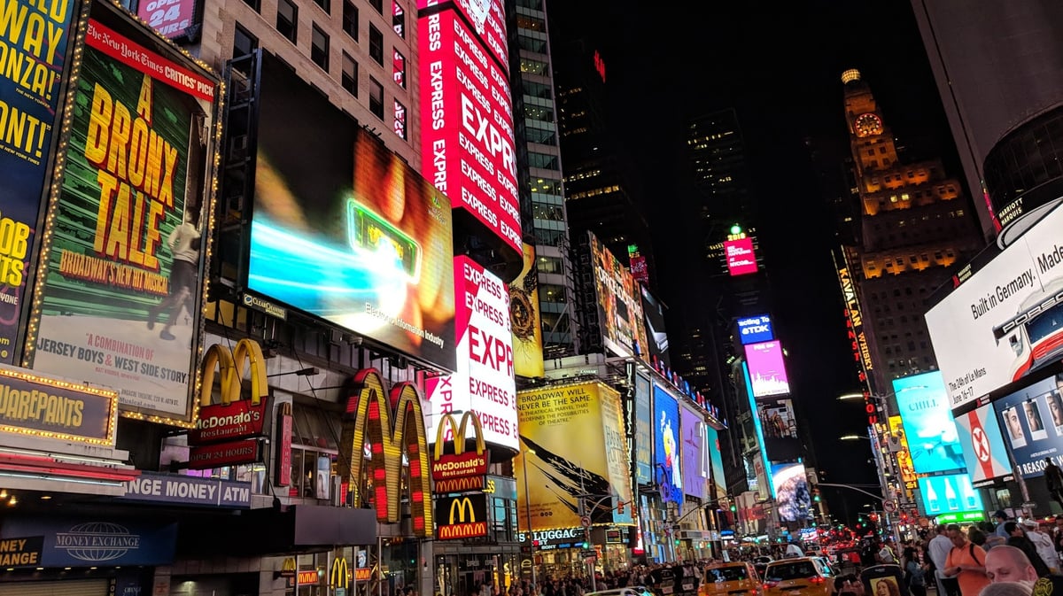 Times Square de noche desde la acera, con las fachadas cubiertas de pantallas gigantes, carteles de musicales de Broadway, los arcos de un McDonald's y los taxis amarillos avanzando entre la gente.