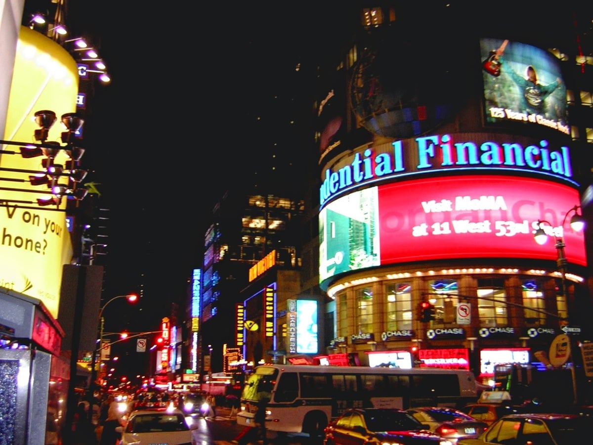 Times Square de noche a principios de los dos mil, con las pantallas y rótulos luminosos de las fachadas encendidos en amarillo, rojo y azul, y el tráfico atascado en la avenida.