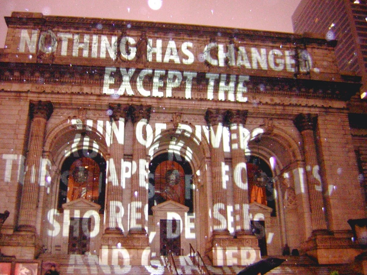 La fachada de mármol blanco de la Biblioteca Pública de Nueva York iluminada de noche, con textos proyectados sobre la piedra y los leones de la entrada en primer plano.