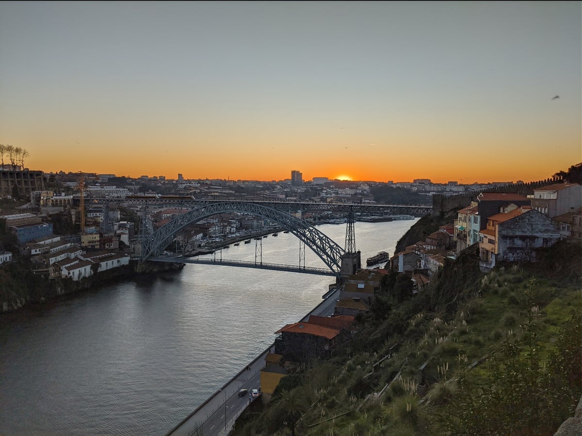 El puente Luis I de Oporto al atardecer, con su gran arco de hierro cruzando el Duero en dos niveles, el sol poniéndose al fondo del río y las casas de Gaia bajando por la ladera de la derecha.