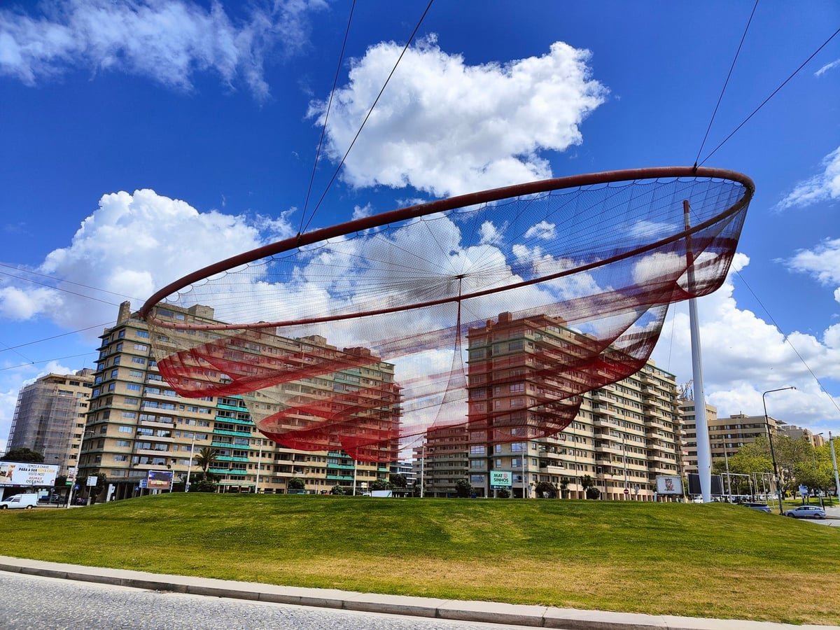 La escultura de red metálica de Janet Echelman suspendida sobre la rotonda del paseo marítimo de Matosinhos, con la playa y el océano Atlántico al fondo.