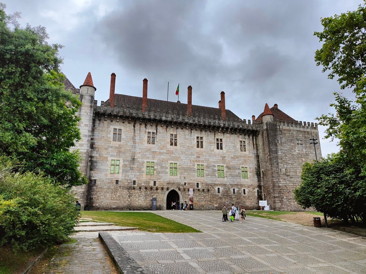 El castillo de Guimarães con sus torres cuadradas de piedra y sus murallas almenadas sobre una elevación de césped, bajo un cielo azul.