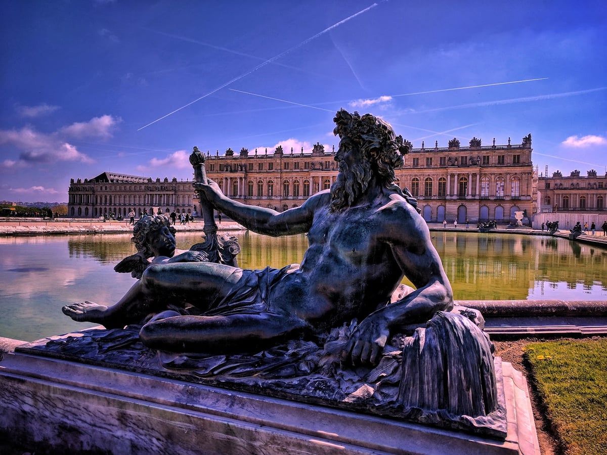 Una escultura de bronce de un dios fluvial recostado, con un amorcillo a su lado, junto al estanque de los jardines de Versalles, con la fachada del palacio extendiéndose entera al fondo bajo un cielo azul con estelas de aviones.