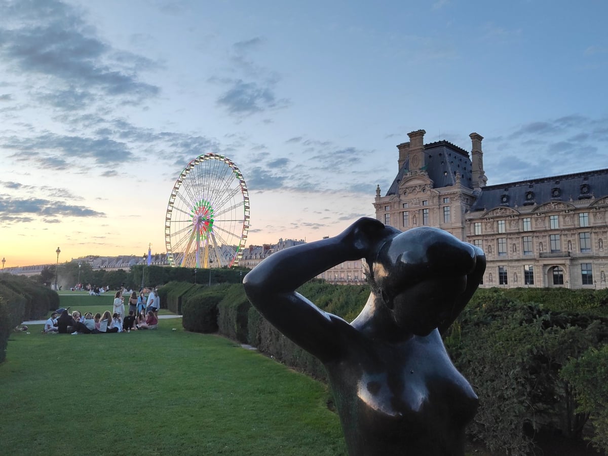 El jardín de las Tullerías al atardecer, con una escultura de bronce de una mujer en primer plano, la noria de la Concordia recortada contra el cielo naranja y el ala del Louvre a la derecha, mientras varios grupos charlan sentados en el césped.