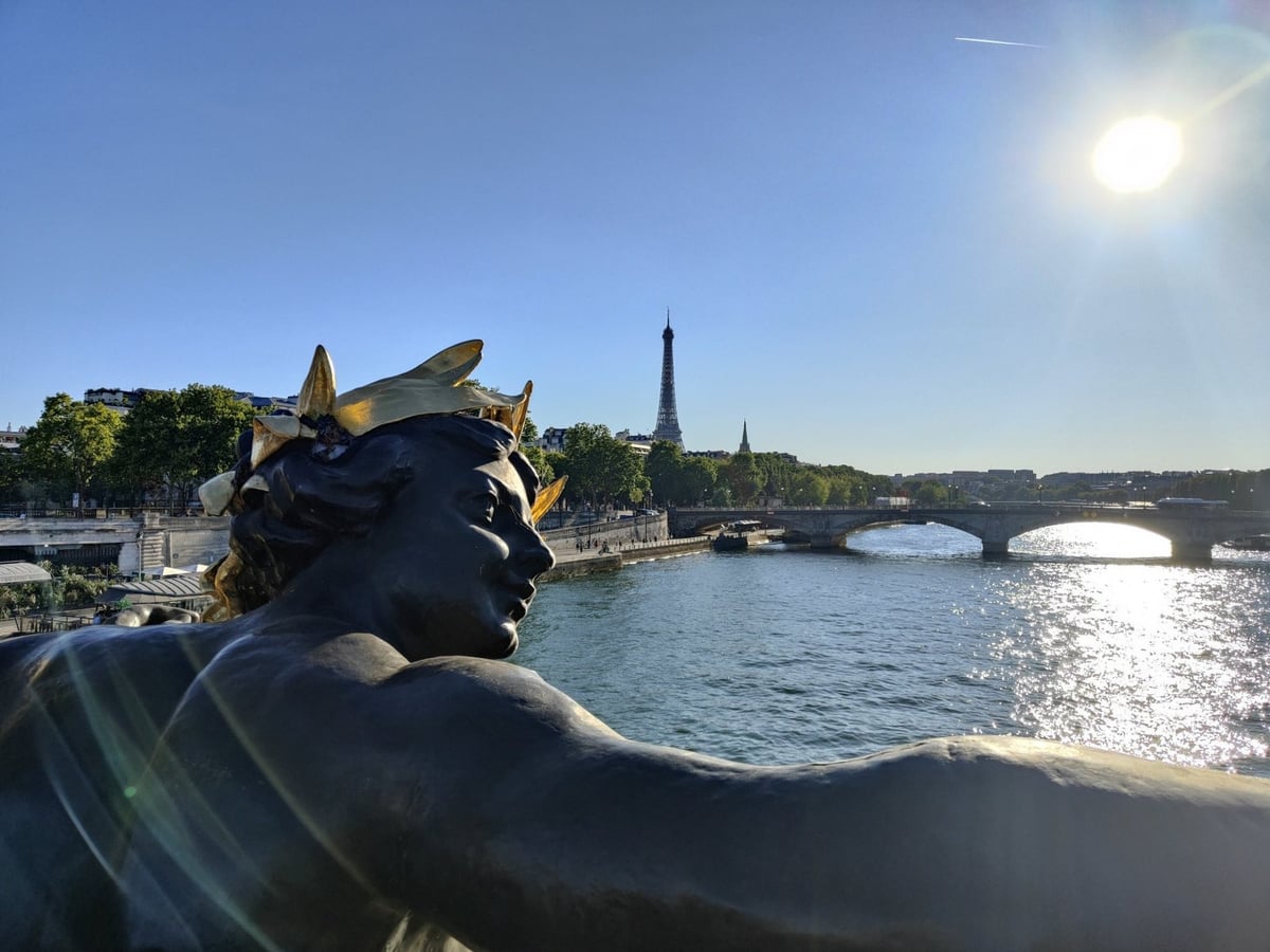 Una de las ninfas de bronce del puente Alejandro III de París vista de cerca, con la corona dorada y el brazo extendido, y detrás el Sena centelleando al sol con la torre Eiffel recortada al fondo.