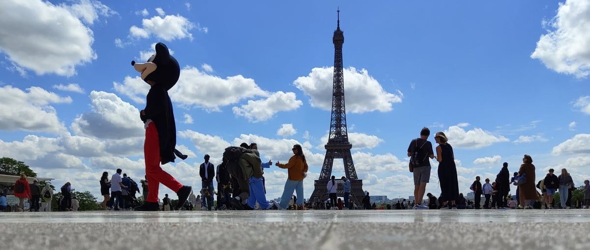 La explanada del Trocadero a ras de suelo, con la torre Eiffel recortada contra un cielo de nubes blancas y, en primer plano, una persona disfrazada de Mickey Mouse cruzando entre los turistas.