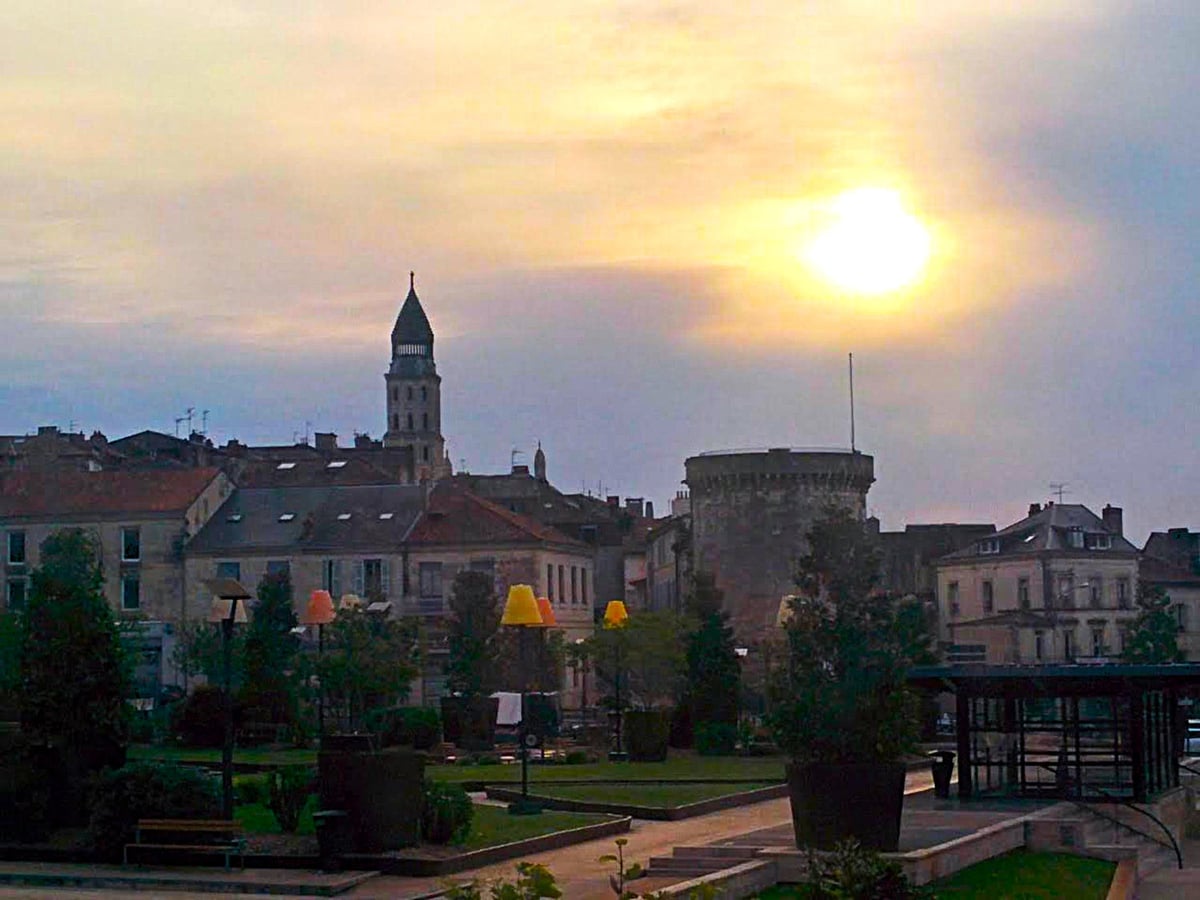 Perigueux al atardecer, con el sol poniéndose entre nubes doradas, el campanario puntiagudo de la iglesia de Saint-Étienne a la izquierda y una torre romana circular a la derecha, sobre los tejados del centro y un jardín en primer plano.