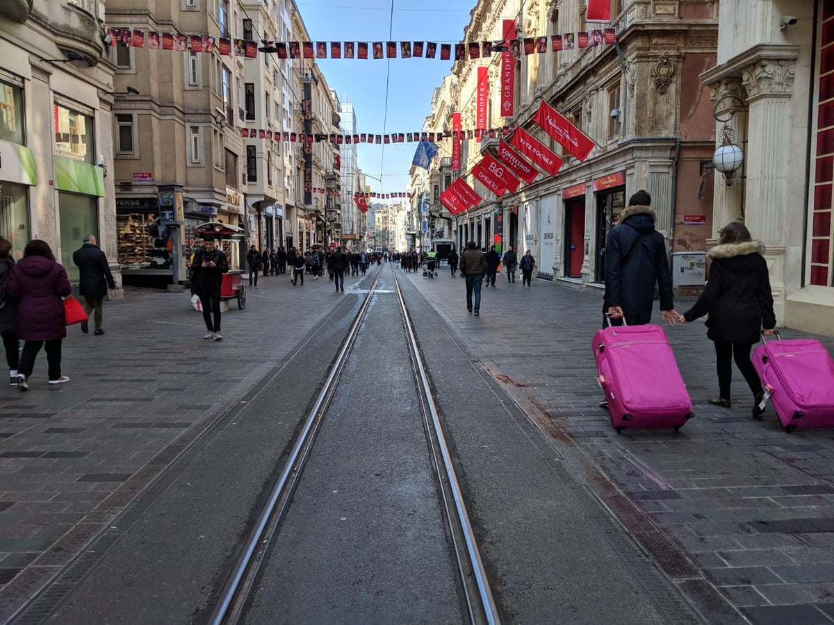 Las vías del tranvía recorren el centro de la avenida İstiklal de Estambul, con guirnaldas de banderines rojos cruzando de lado a lado y una pareja arrastrando dos maletas rosas por la acera de la derecha.
