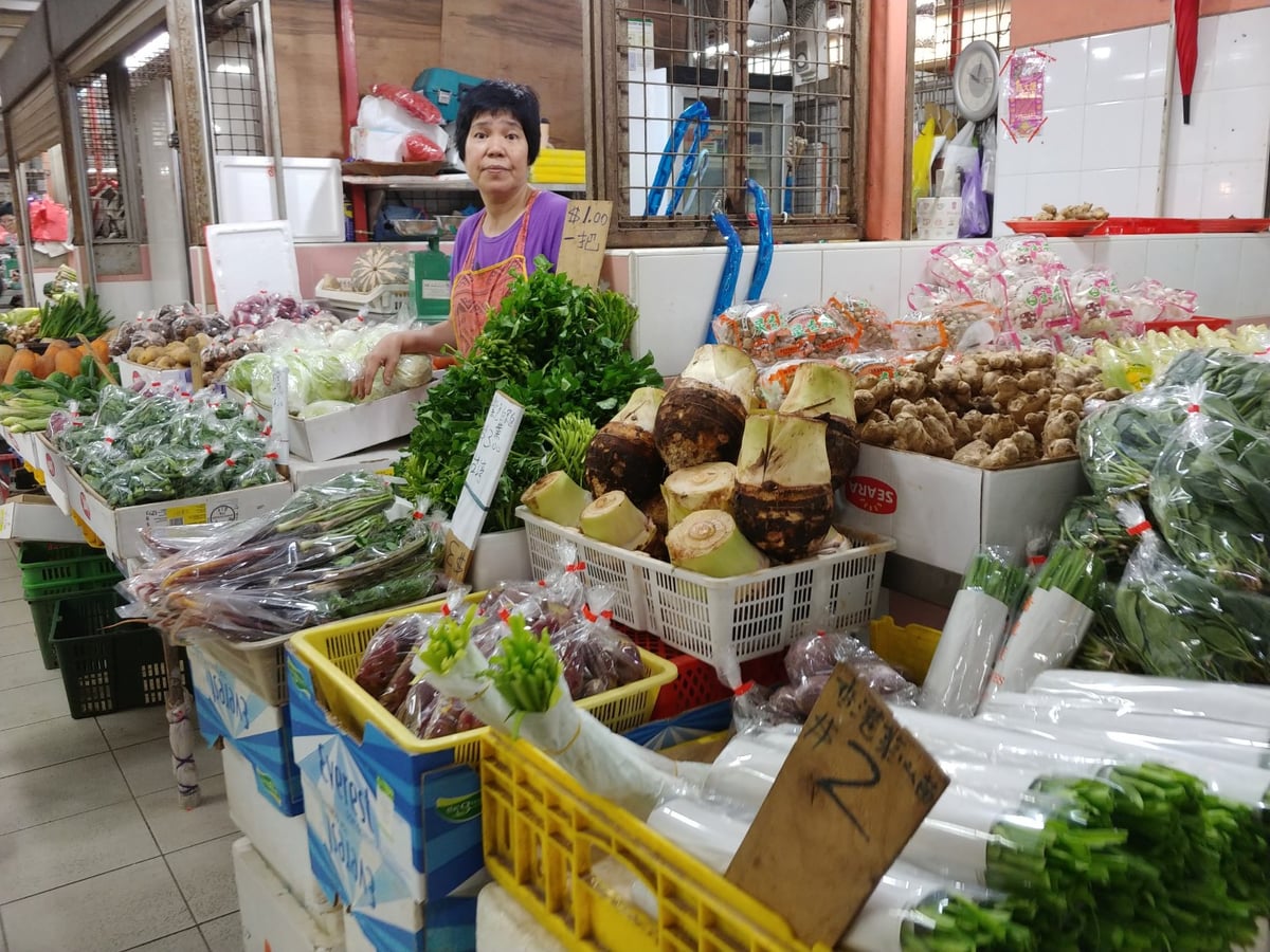 Una vendedora mira a la cámara desde detrás del mostrador de su puesto de verduras en un mercado asiático, rodeada de cajas de hortalizas, raíces de taro y manojos de hierbas con los precios escritos a mano.