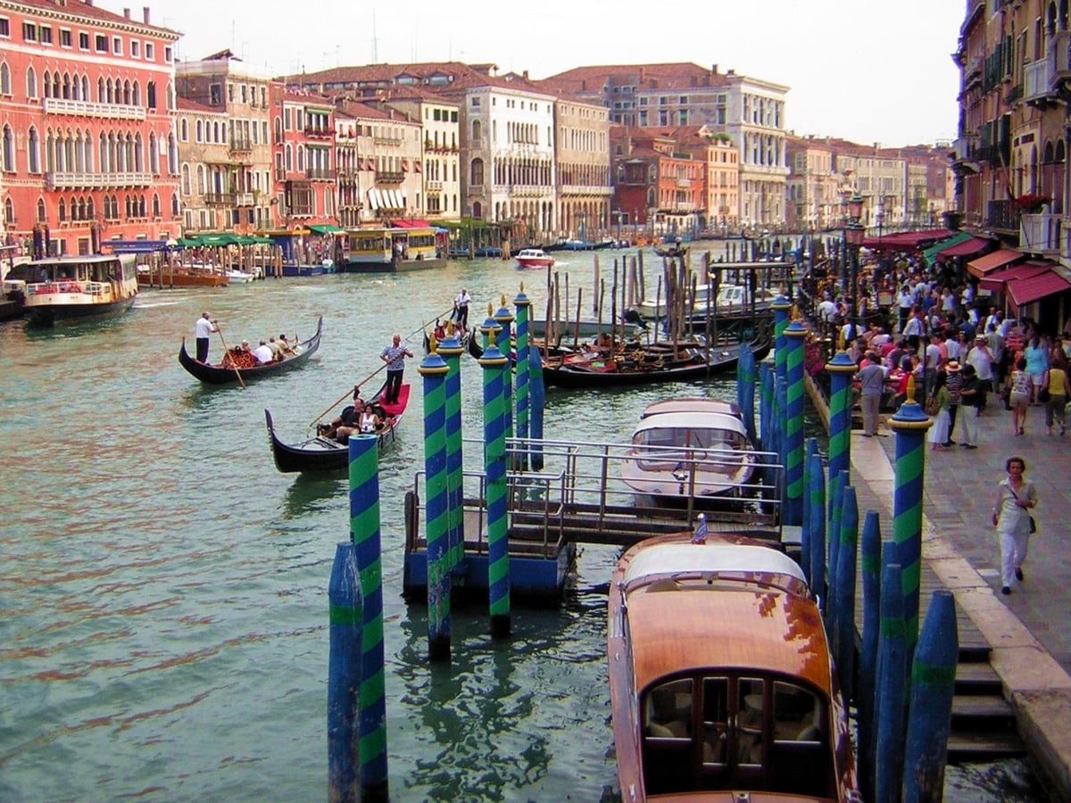 El Gran Canal de Venecia con dos góndolas cargadas de turistas, los postes de amarre pintados a rayas azules y verdes en primer plano, los palacios alineados en la orilla y una multitud abarrotando el muelle de la derecha.