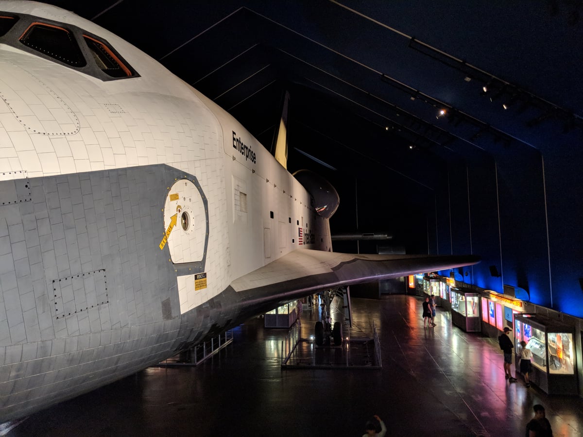 El transbordador espacial Enterprise expuesto en la penumbra de un museo, con el morro blanco recortado en primer plano, el ala extendida sobre la sala y unos visitantes diminutos junto a las vitrinas del fondo.