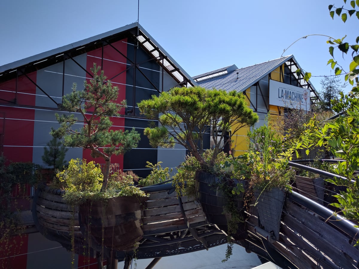 Una pasarela de acero con grandes jardineras de madera plantadas con pinos y arbustos, montada delante de una nave industrial pintada a franjas rojas, grises y amarillas con el rótulo «La Machine».