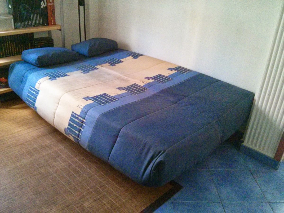 Un sofá cama azul abierto en el salón de una casa particular, hecho con una colcha de rayas, dos cojines a modo de almohada y una estantería de libros justo detrás.