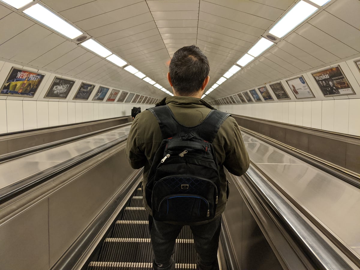 Un hombre con mochila al hombro baja de espaldas por la escalera mecánica de una estación de metro, en un túnel largo forrado de azulejos blancos y carteles de espectáculos a ambos lados.