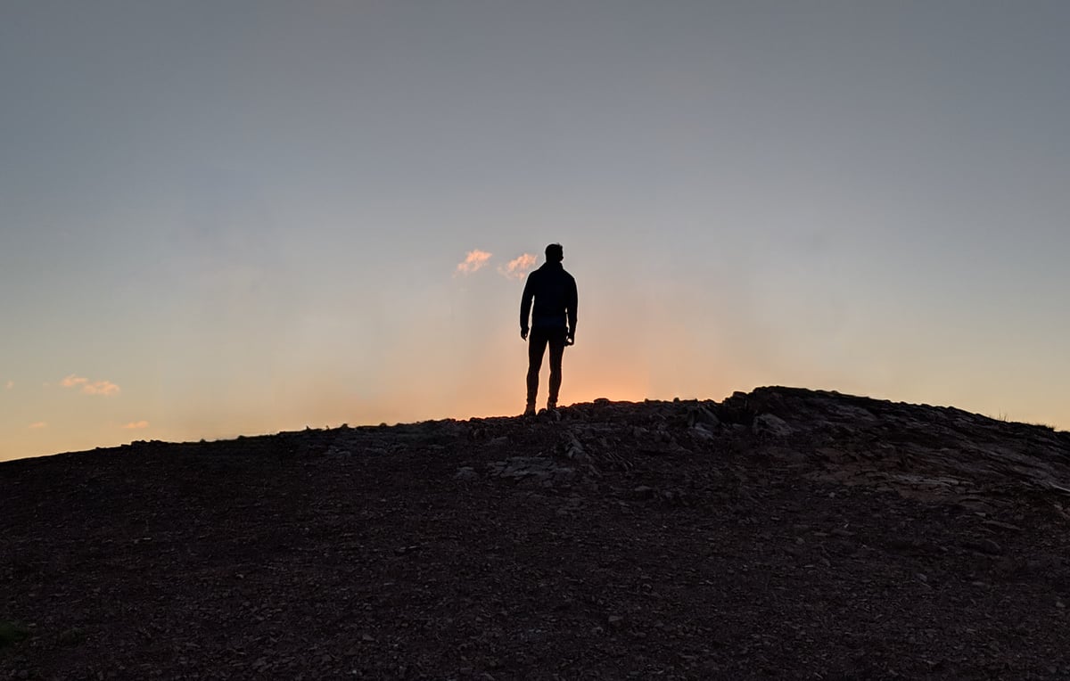 Silueta de una persona sola de pie en lo alto de una loma pedregosa, recortada contra el cielo naranja y azul del atardecer.