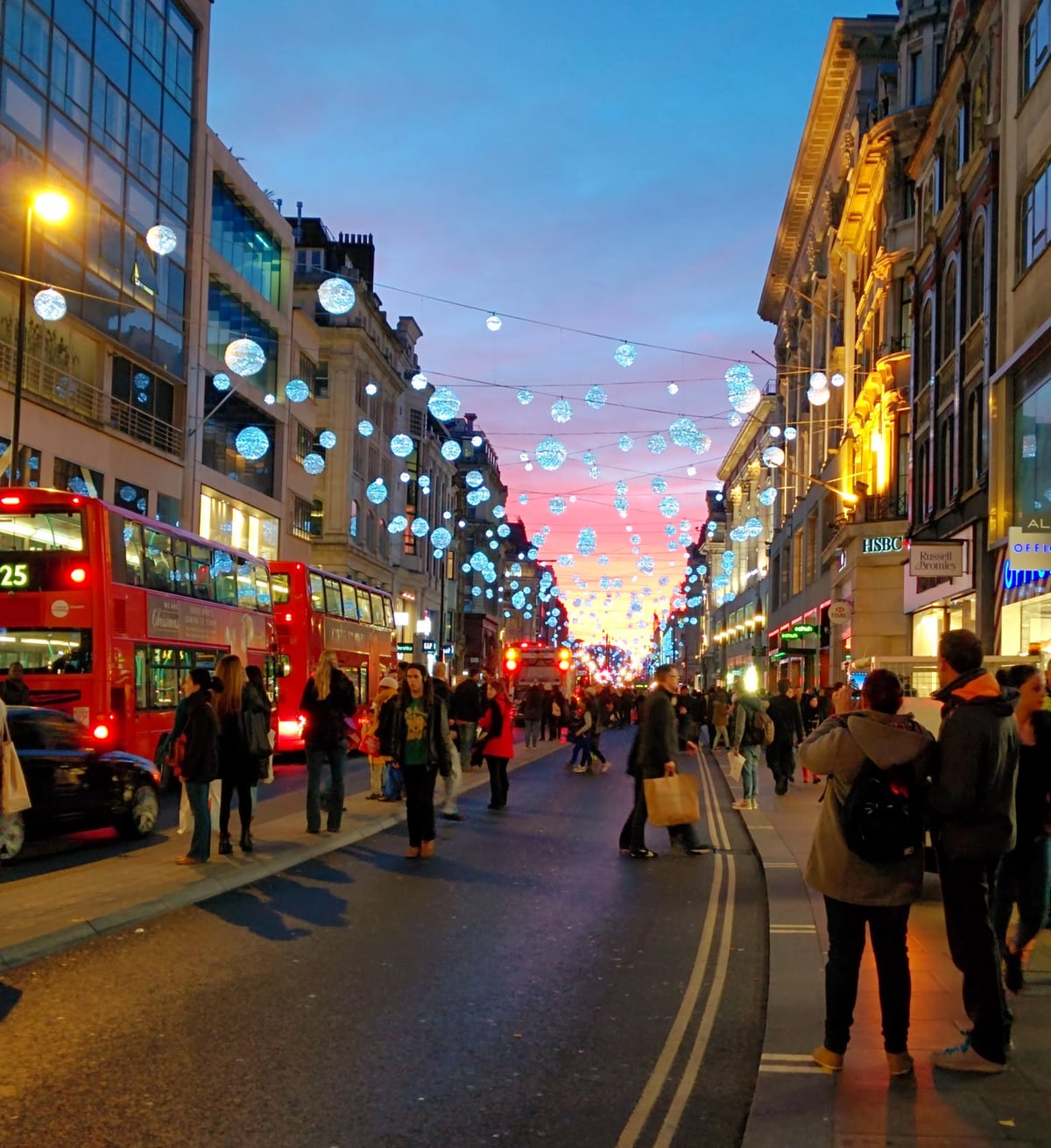 Oxford Street en Navidad al anochecer, con cientos de bolas luminosas colgadas de un lado a otro de la calle, dos autobuses rojos de dos pisos a la izquierda y la gente cargada de bolsas bajo un cielo rosado.