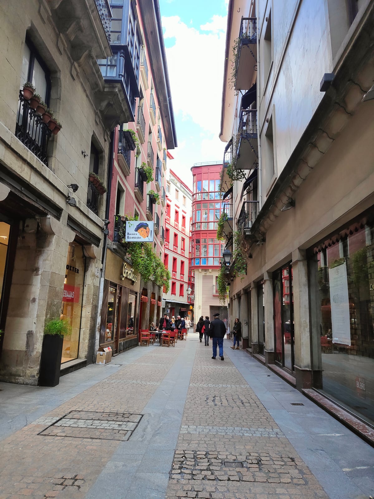 Calle estrecha del Casco Viejo de Bilbao con soportales de piedra, miradores acristalados de colores y un edificio rojo cerrando el fondo, con las mesas de una terraza a un lado y unos pocos paseantes.