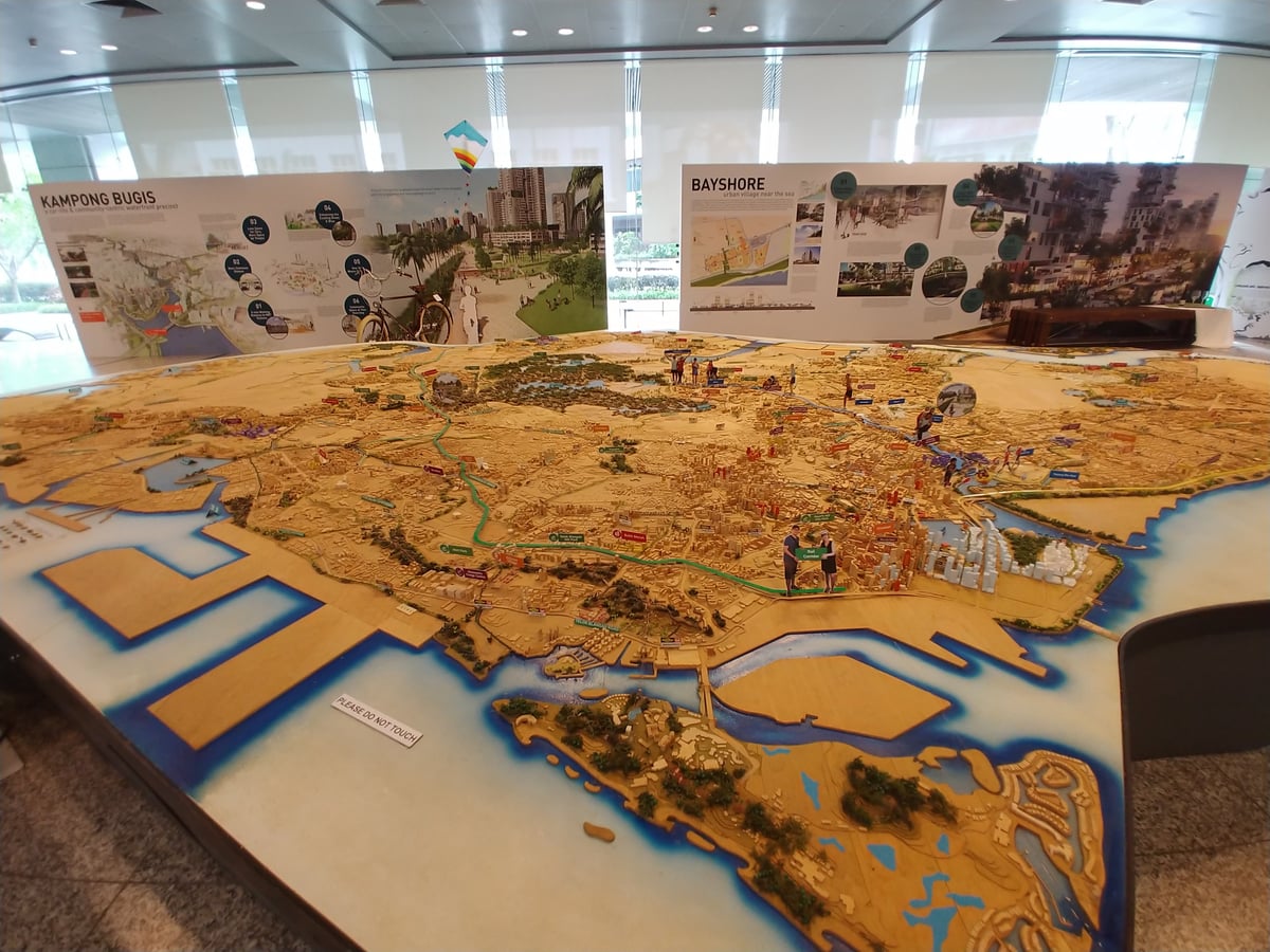 Gran maqueta de Singapur que ocupa una sala entera, con la isla reproducida edificio a edificio en madera clara, el mar en azul y varios visitantes inclinados sobre ella al fondo.