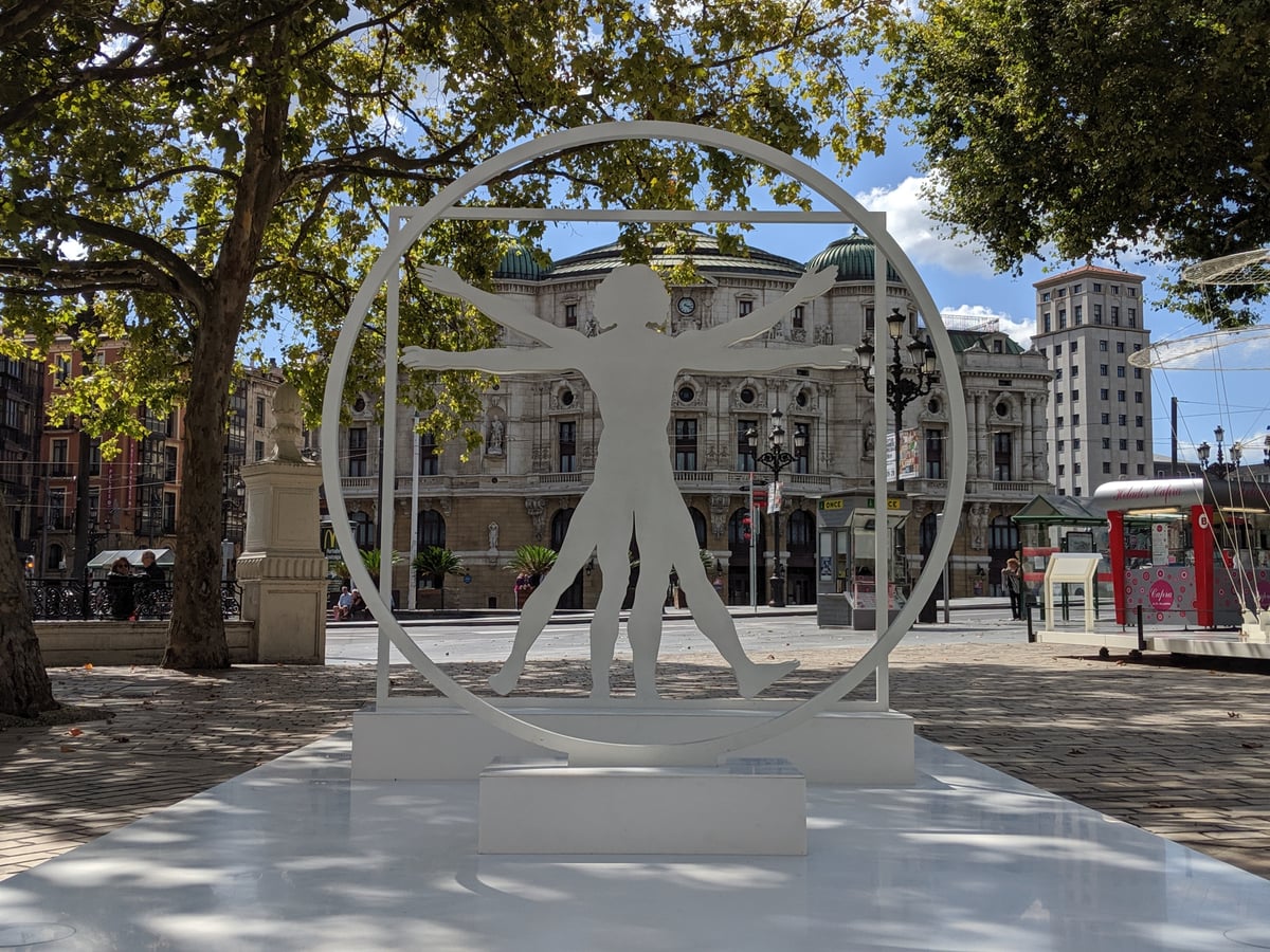 Escultura blanca del Hombre de Vitruvio, recortada en chapa dentro de un círculo y un cuadrado, instalada en el paseo de El Arenal de Bilbao con la fachada del Teatro Arriaga justo detrás.
