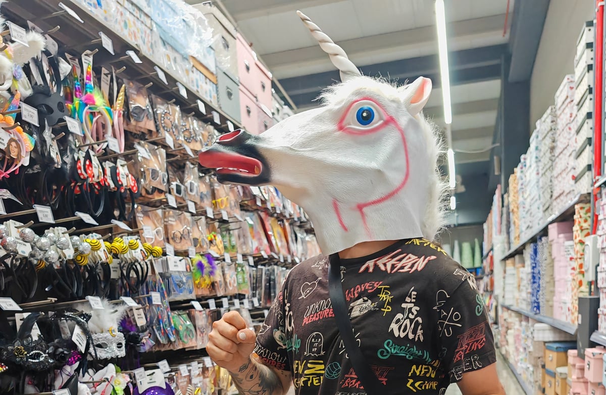 Una persona con una máscara de goma de unicornio blanco, de boca abierta y crin de pelo, posa en el pasillo de una tienda de disfraces entre estanterías repletas de diademas, antifaces y complementos.