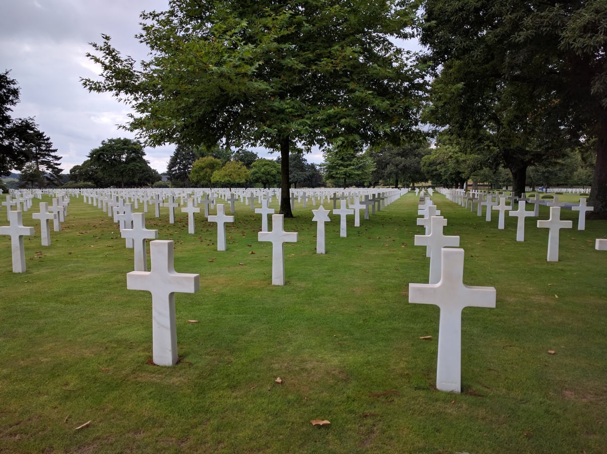 Filas y filas de cruces de mármol blanco alineadas sobre el césped de un cementerio militar estadounidense, con una estrella de David entre ellas y grandes árboles dando sombra al fondo.