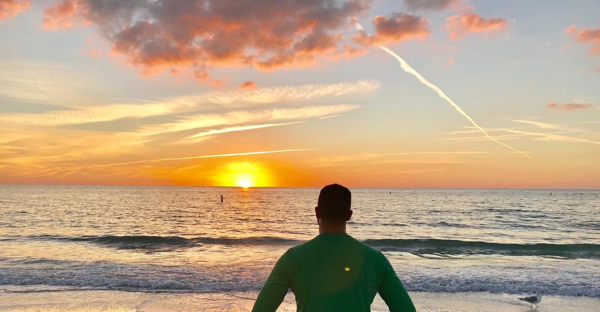 Una persona de espaldas, con jersey verde, mira el atardecer desde la orilla, con el sol tocando el horizonte del mar, las nubes teñidas de naranja y una gaviota en la arena a la derecha.