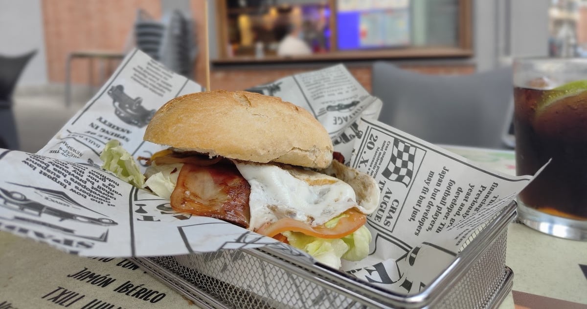 Un bocadillo abierto de bacon, huevo frito, tomate y lechuga servido en una cestita metálica sobre papel de periódico, en la mesa de una terraza, con un refresco con hielo al lado.