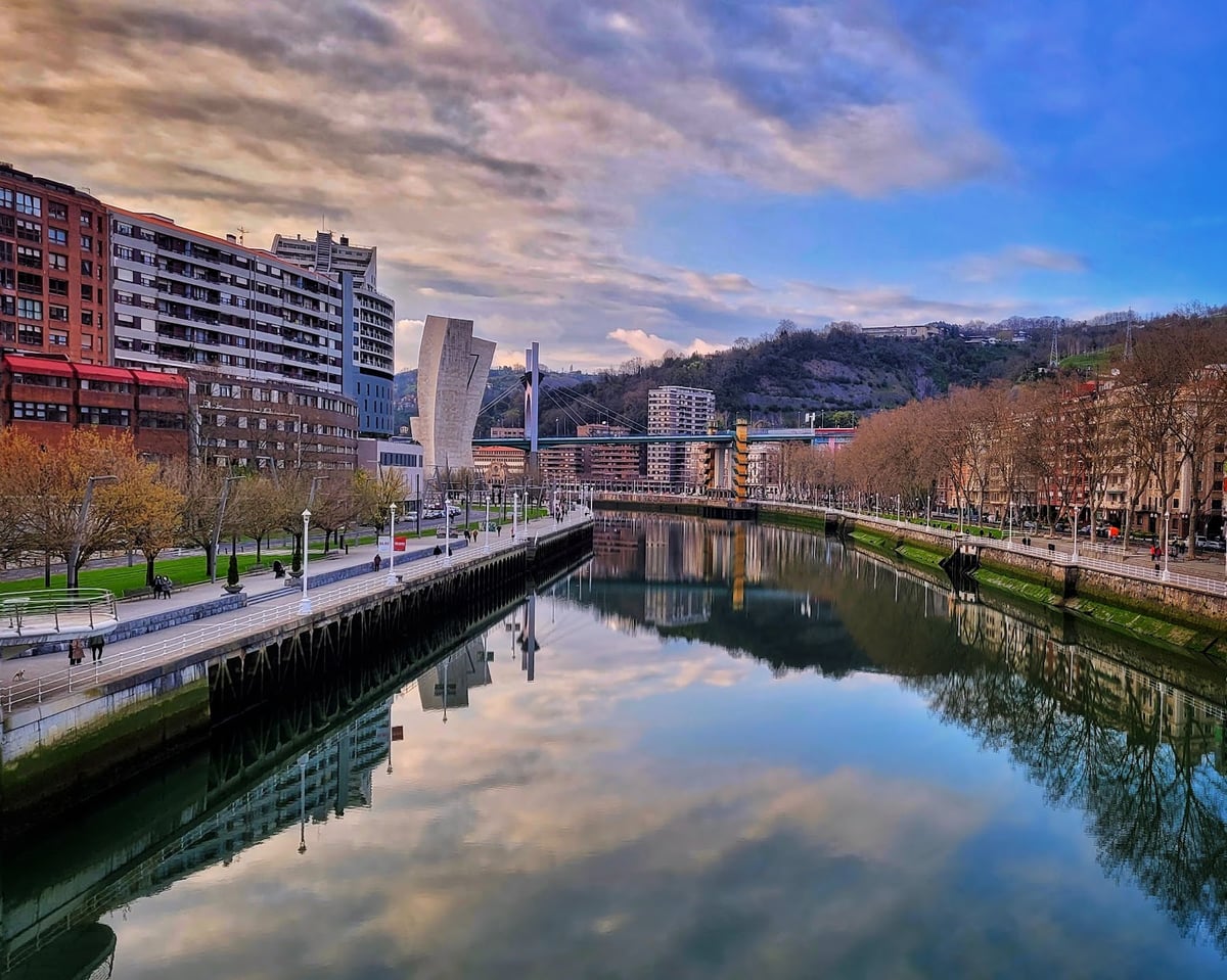 La ría de Bilbao al atardecer, con el agua completamente en calma reflejando las nubes rosadas, los paseos arbolados a ambas orillas y la torre del Museo Marítimo asomando entre los edificios.