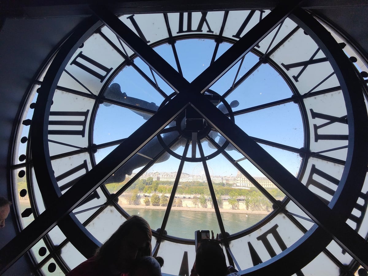 Vista desde detrás del gran reloj del Museo de Orsay, con los números romanos y la maquinaria recortados a contraluz y, al otro lado del cristal, el Sena y los tejados de París.