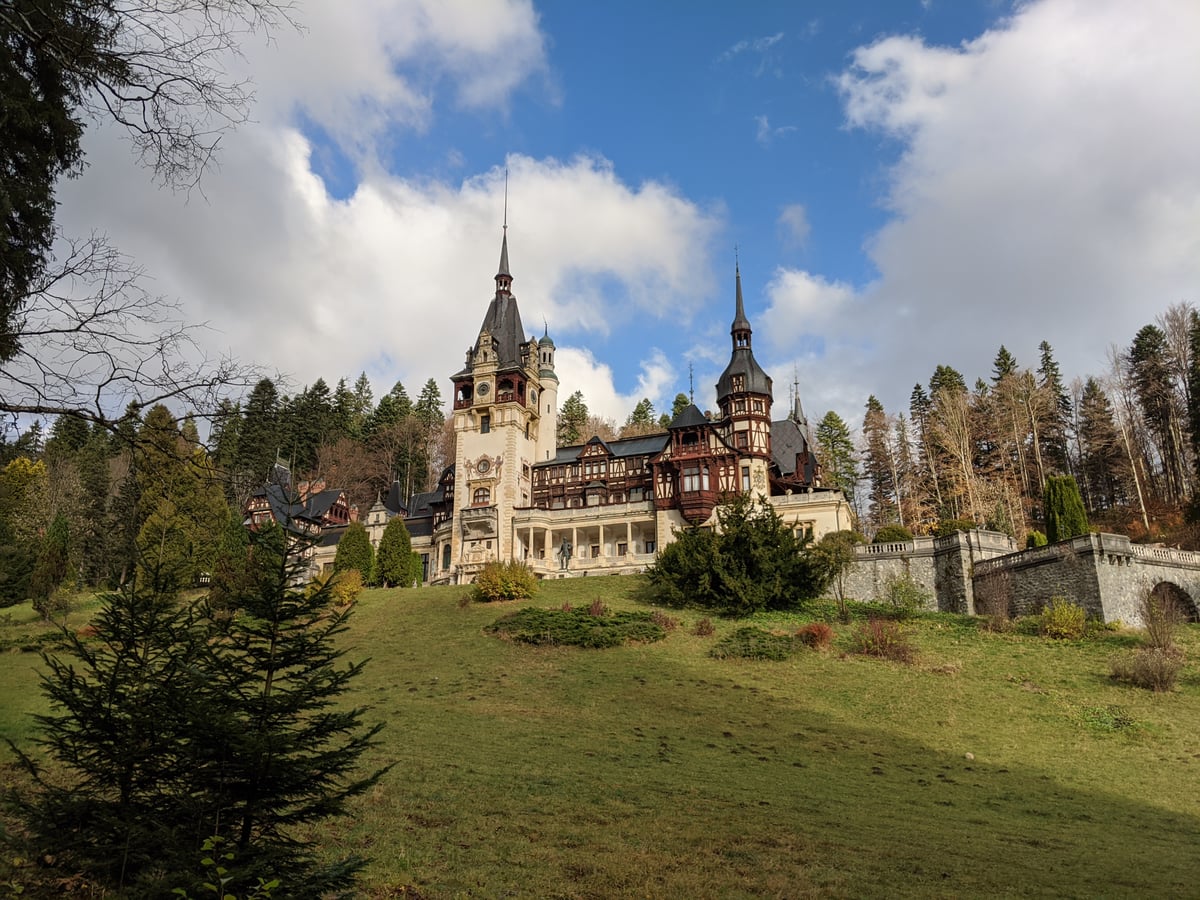 El castillo de Peleș en Rumanía, con sus torres puntiagudas, los entramados de madera y la fachada clara, encaramado sobre una pradera en cuesta y rodeado de abetos, bajo un cielo de nubes blancas