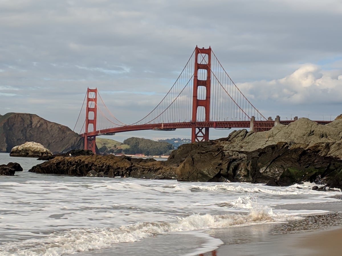 El puente Golden Gate visto desde la orilla de Baker Beach, con las torres rojas recortadas contra un cielo nublado, las rocas oscuras de la playa en primer plano y la espuma de una ola llegando a la arena.