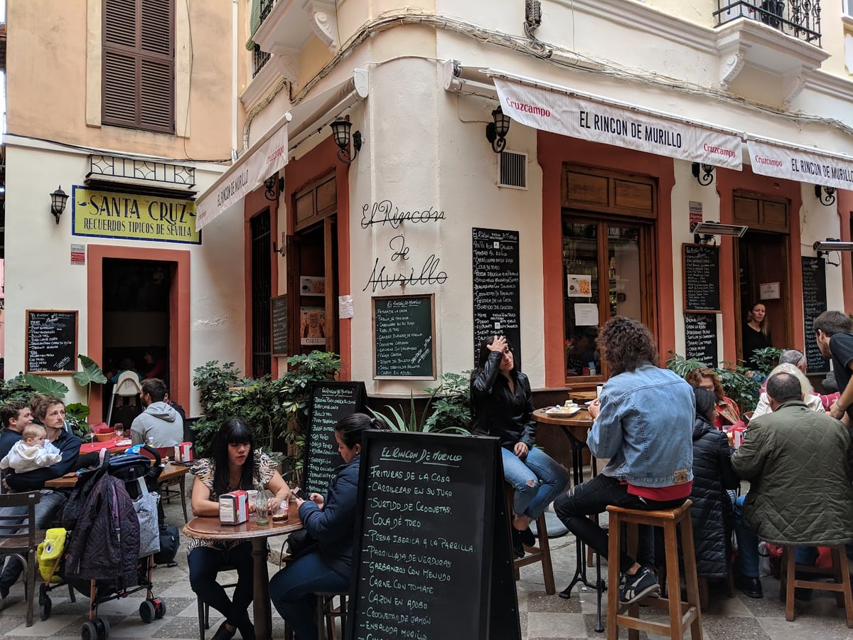 La terraza del bar El Rincón de Murillo, en el barrio de Santa Cruz de Sevilla, llena de gente en mesas altas y taburetes, con las pizarras de tapas escritas a mano y una tienda de recuerdos en la esquina.