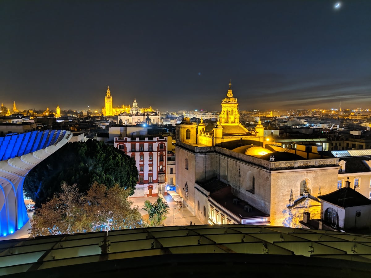 Sevilla de noche desde lo alto de las Setas, con la Giralda iluminada al fondo, la torre barroca de la iglesia de la Anunciación en primer plano y la estructura de madera de la pasarela cruzando la imagen.