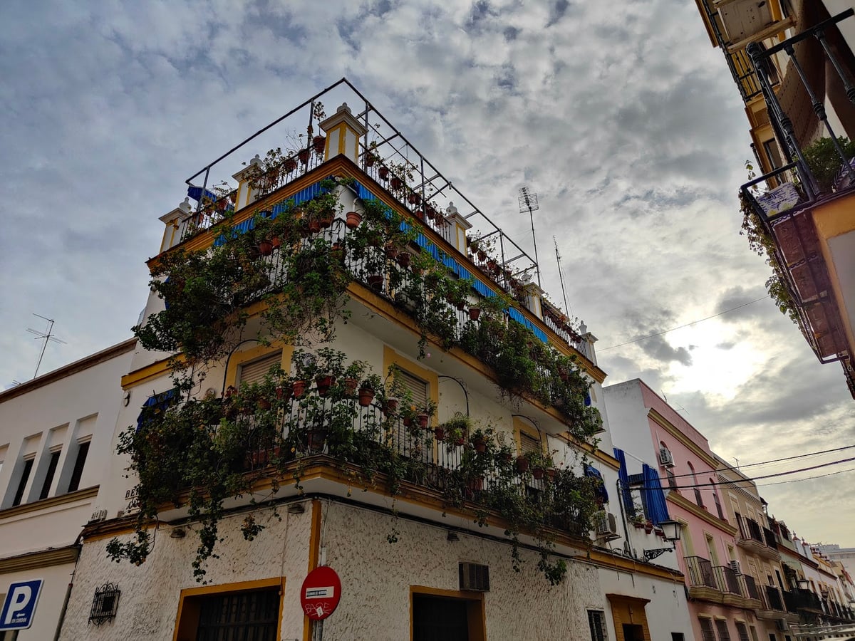 Un edificio de Triana con las balconadas cubiertas de macetas y plantas colgantes, bajo un cielo nuboso