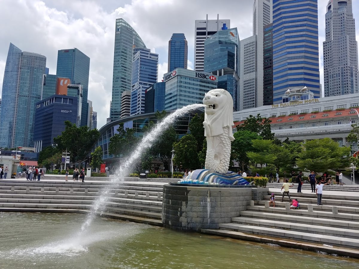 La fuente del Merlión de Singapur lanzando su chorro de agua a la bahía, con las torres del distrito financiero al fondo y el edificio colonial del Fullerton Hotel a la derecha.