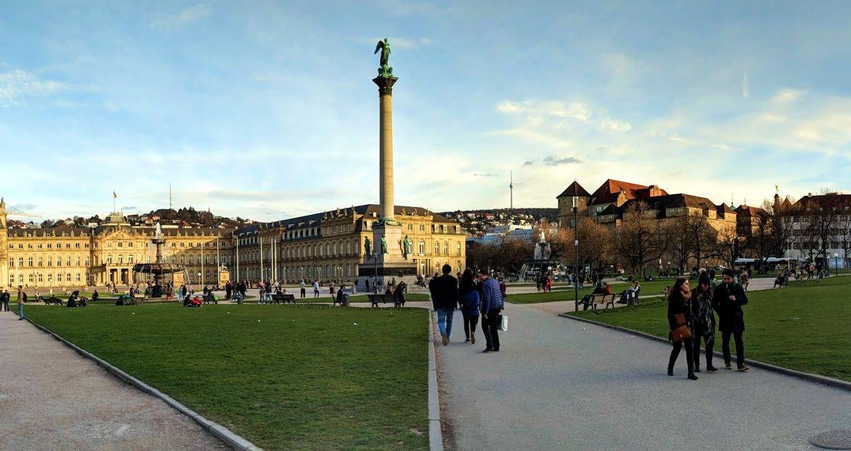 La plaza del Palacio de Stuttgart al atardecer, con la columna del Jubileo coronada por una figura dorada en el centro, el Palacio Nuevo a la izquierda, el césped y la gente paseando por los caminos.