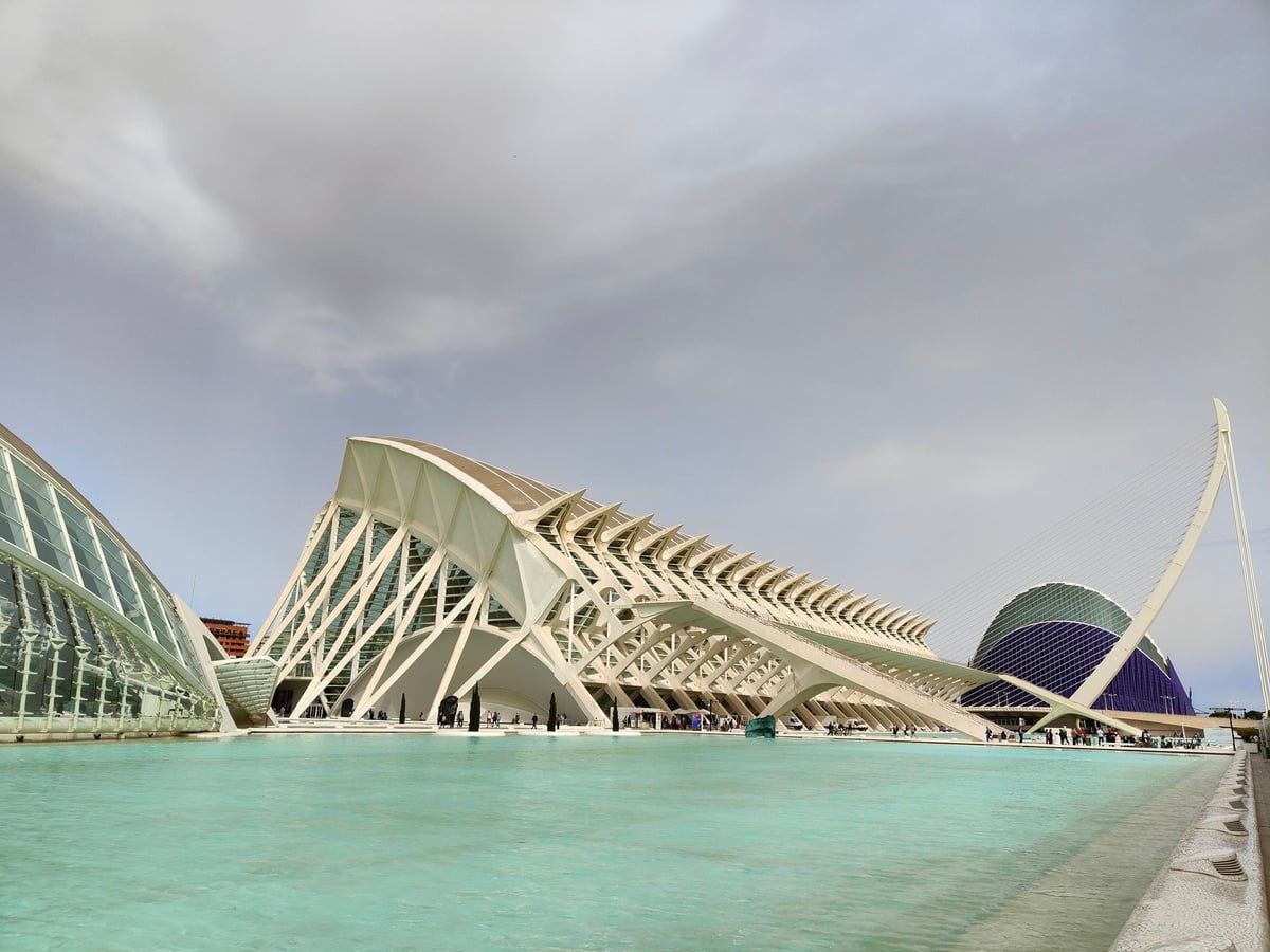La Ciudad de las Artes y las Ciencias de Valencia reflejada en el estanque turquesa, con el esqueleto blanco del Museo de las Ciencias en el centro, el Hemisfèric a la izquierda y el Ágora azul al fondo.