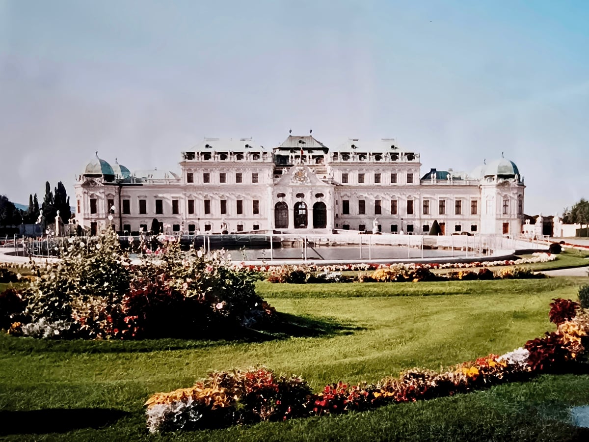 El palacio del Belvedere de Viena en una foto de carrete, con la fachada barroca blanca y sus cúpulas verdes reflejándose en el estanque, y los parterres de flores del jardín en primer plano.