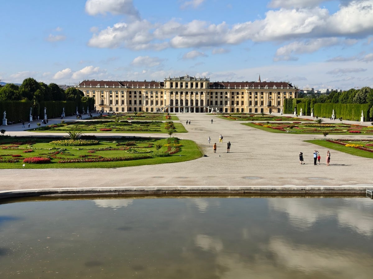 El palacio de Schönbrunn de Viena visto desde los jardines, con su larga fachada amarilla al fondo, los parterres de flores simétricos, la gran explanada de grava y el estanque en primer plano.