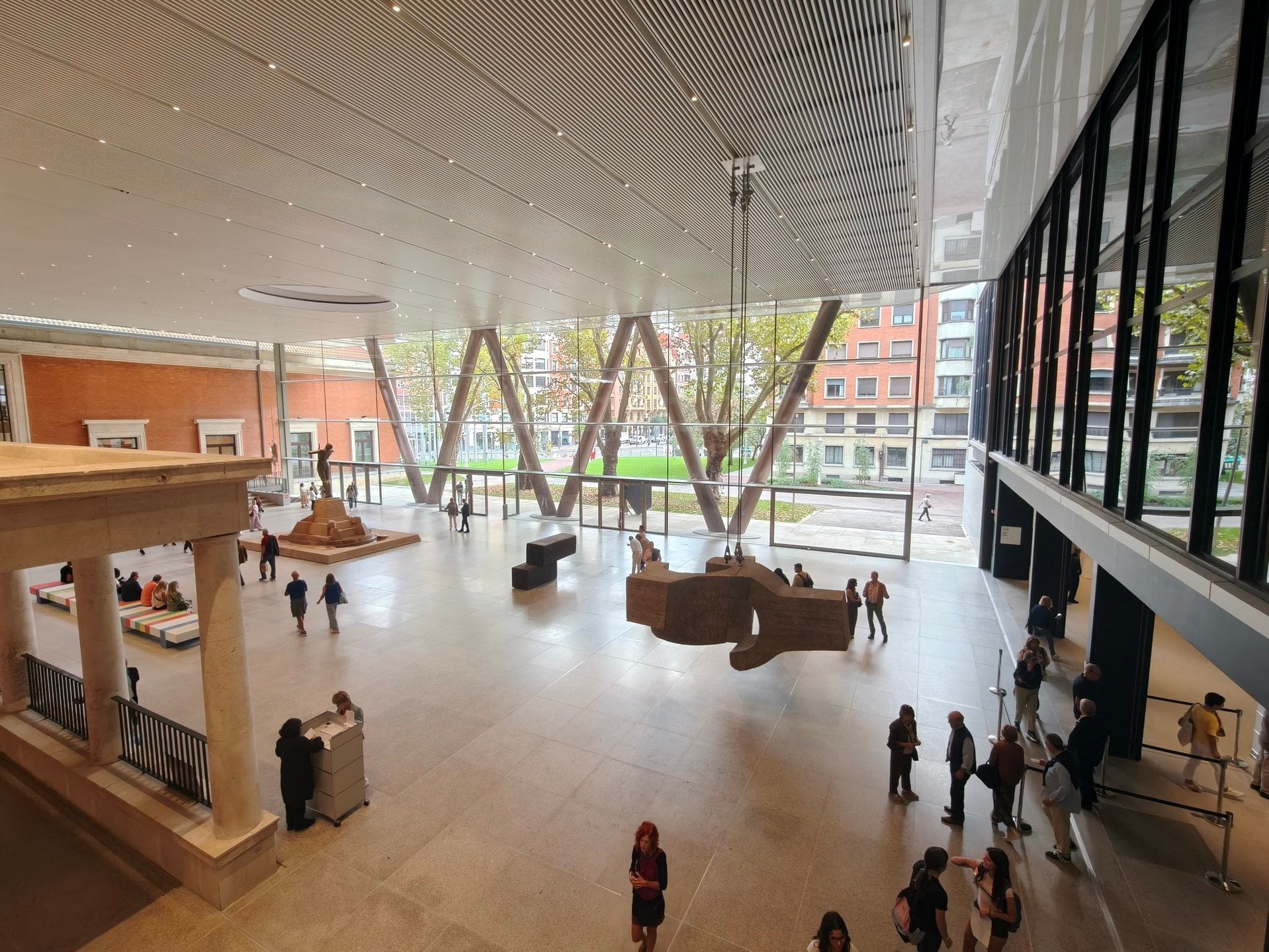 Atrio Arriaga del Museo de Bellas Artes de Bilbao visto desde arriba, con los pilares en V de la nueva estructura tras el cristal, la fuente de Durrio al fondo y visitantes bajo una escultura suspendida