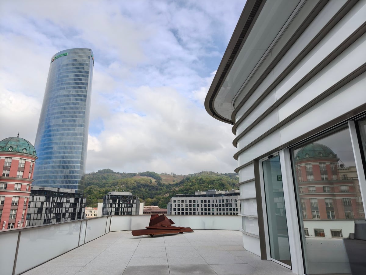Terraza de Agravitas con una escultura de acero oxidado, la Torre Iberdrola al fondo y los paneles blancos de la nueva piel exterior a la derecha