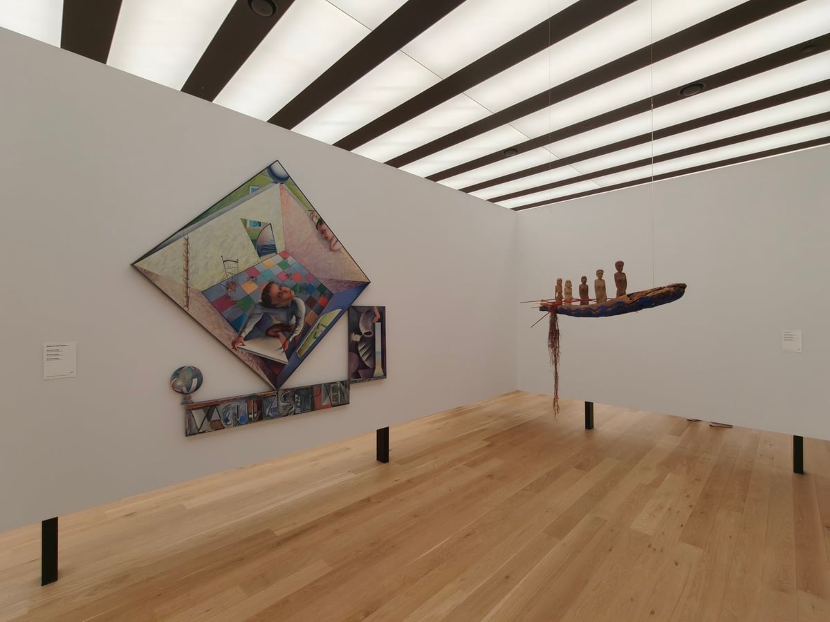 Sala del edificio de los años setenta con tarima clara, techo de franjas luminosas, una pintura en forma de rombo en una pared y una barca con figuras suspendida