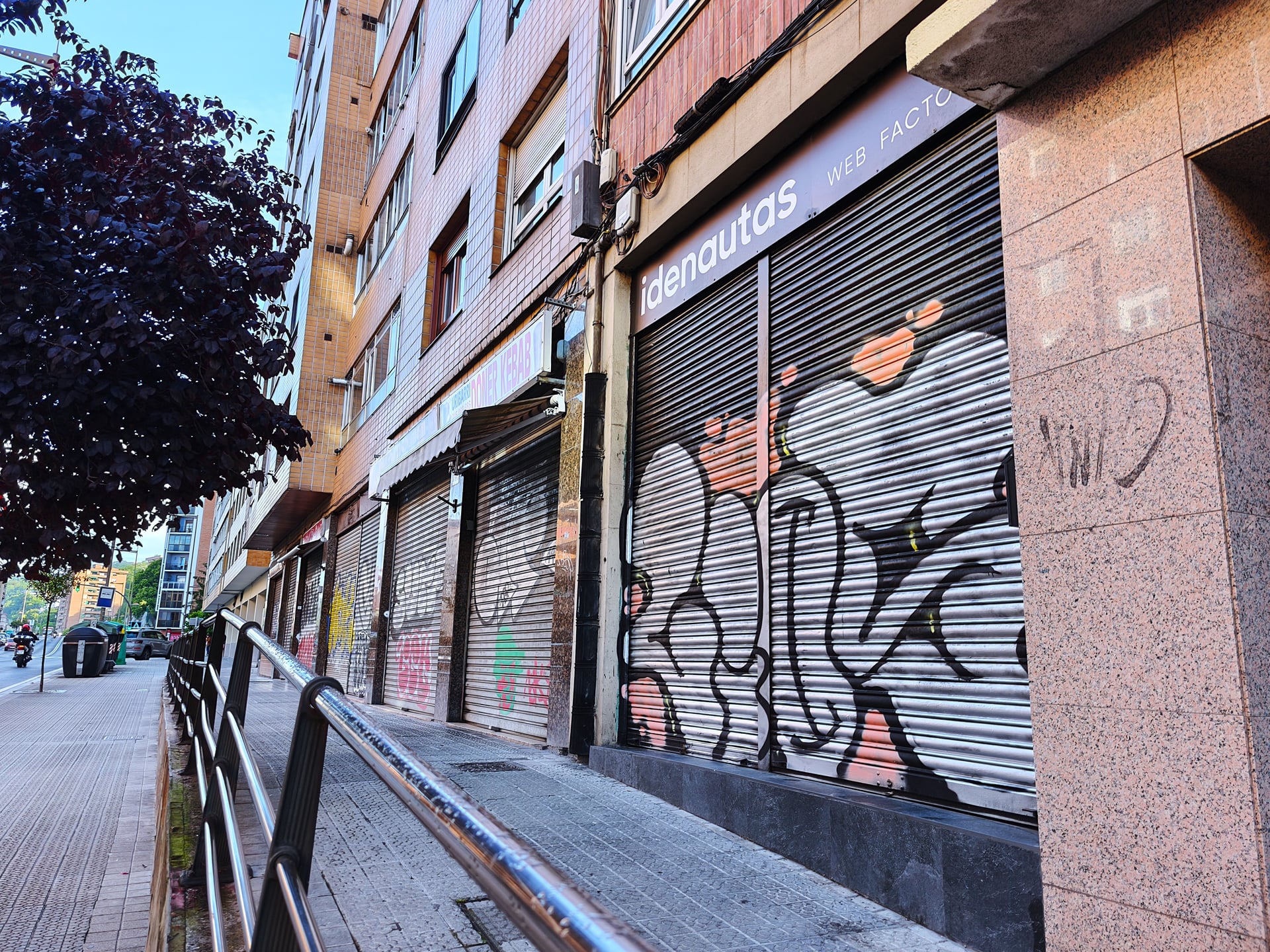 Fila de persianas metálicas cerradas en una calle urbana, cubiertas de grafitis y tags, iluminadas por una farola en la penumbra del amanecer